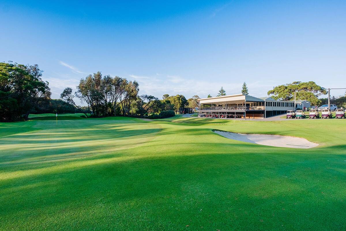 View of the 18th hole fairway and green at Shoalhaven Heads Golf Club, featuring manicured green fairways, sand bunkers, and the clubhouse nestled among mature trees under a clear blue sky.