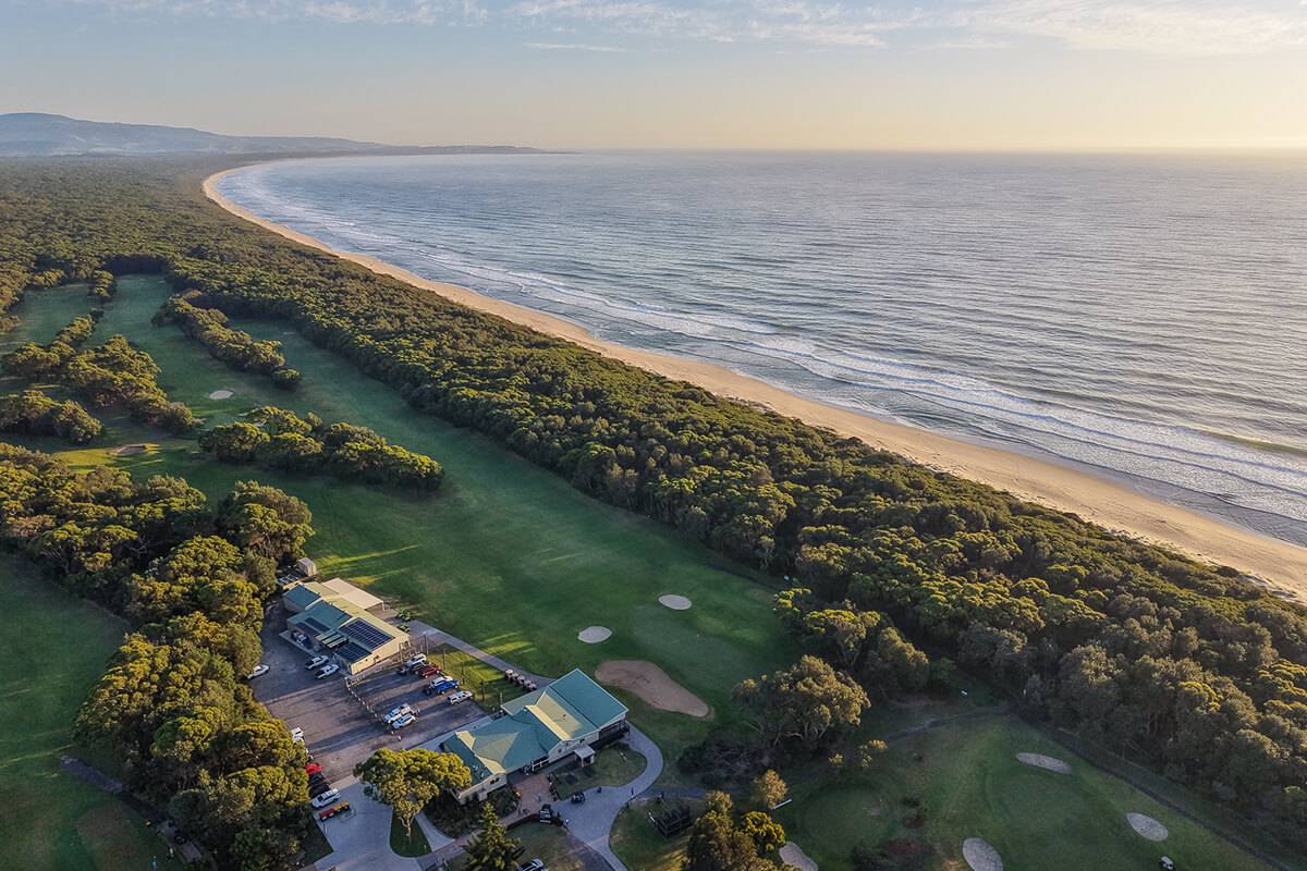 Aerial view of Shoalhaven Heads Golf Club and clubhouse nestled between lush green fairways and a sweeping sandy beach along the coastline, surrounded by native bushland at golden hour.